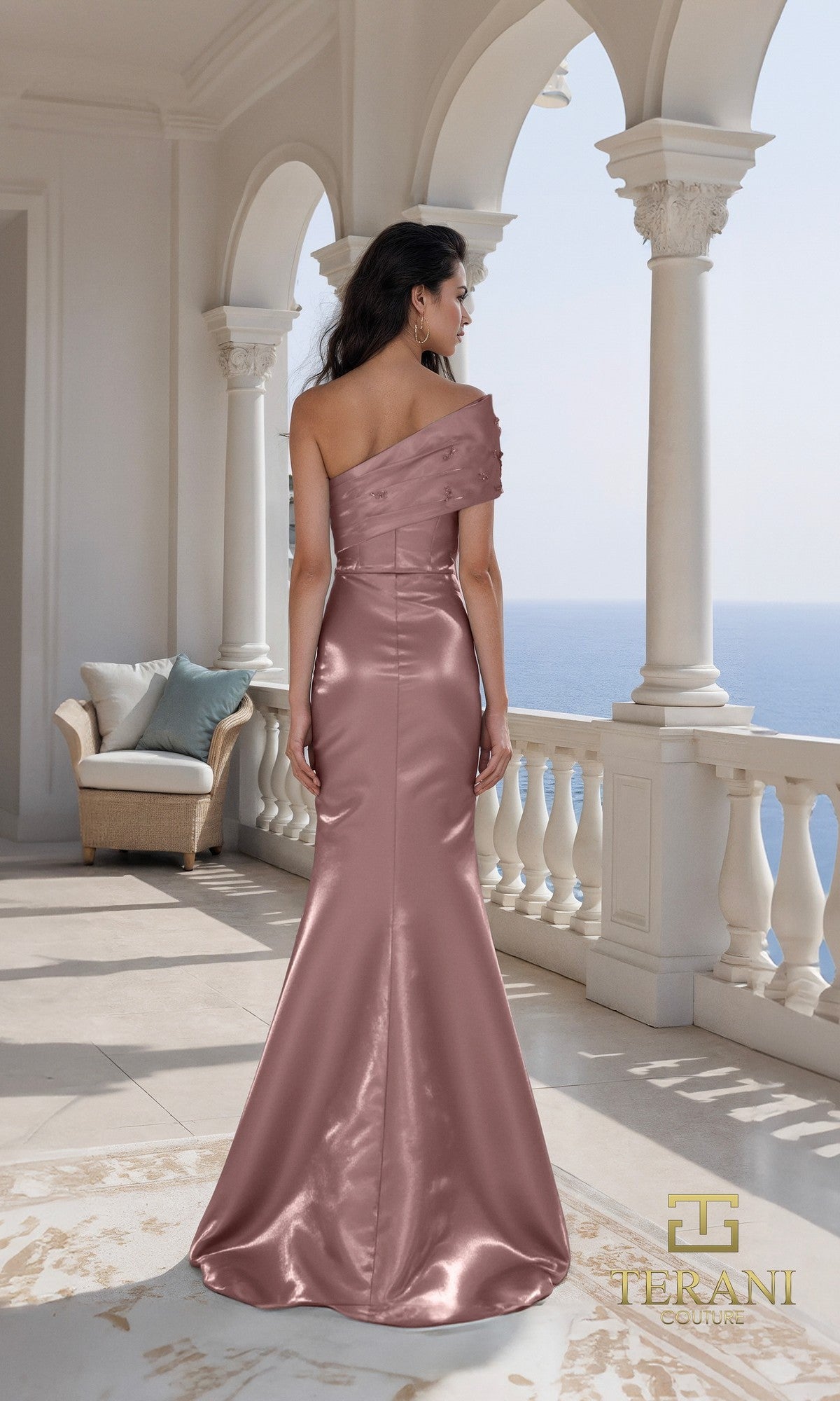 Woman in a Terani 261E6227 long formal dress stands on a balcony overlooking the ocean, with white columns and a wicker chair in the background.